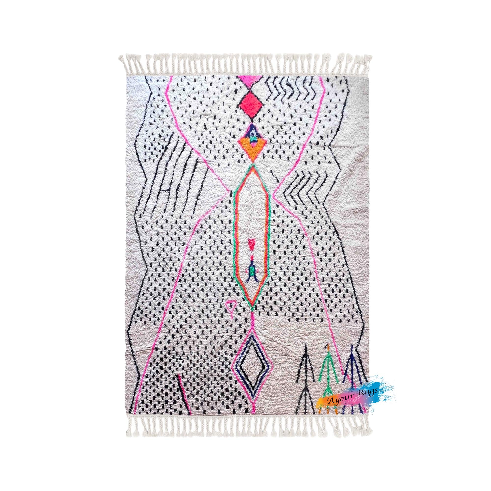 a vibrant, abstract rug with a geometric pattern in shades of pink, black, and white. The rug has a fringe at the bottom and is displayed against a white background.